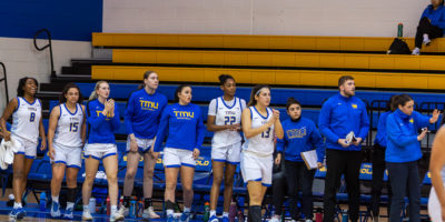 The TMU Bold women's basketball team from the bench standing and paying attention to the game