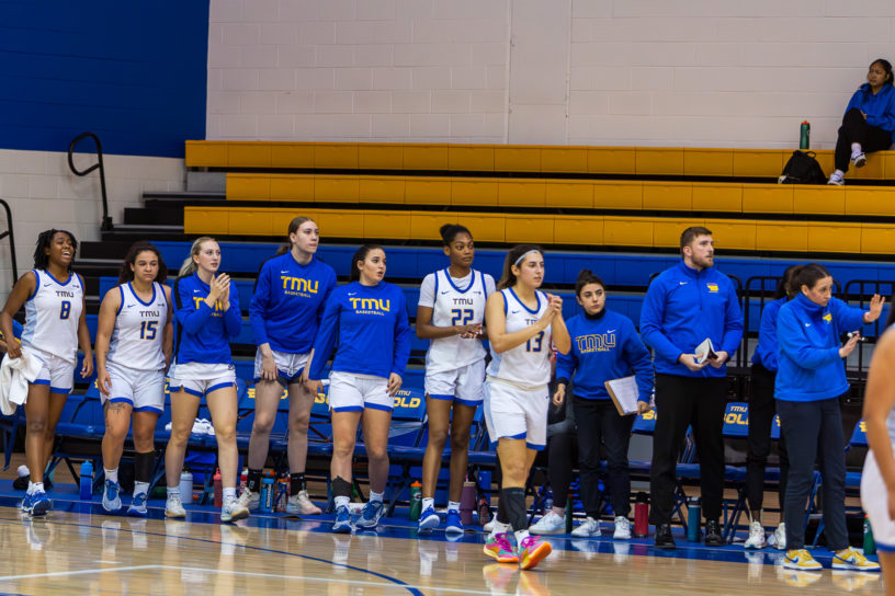 The TMU Bold women's basketball team from the bench standing and paying attention to the game