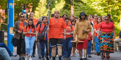 A group of people in orange shirts walk mournfuly down the TMU campus.