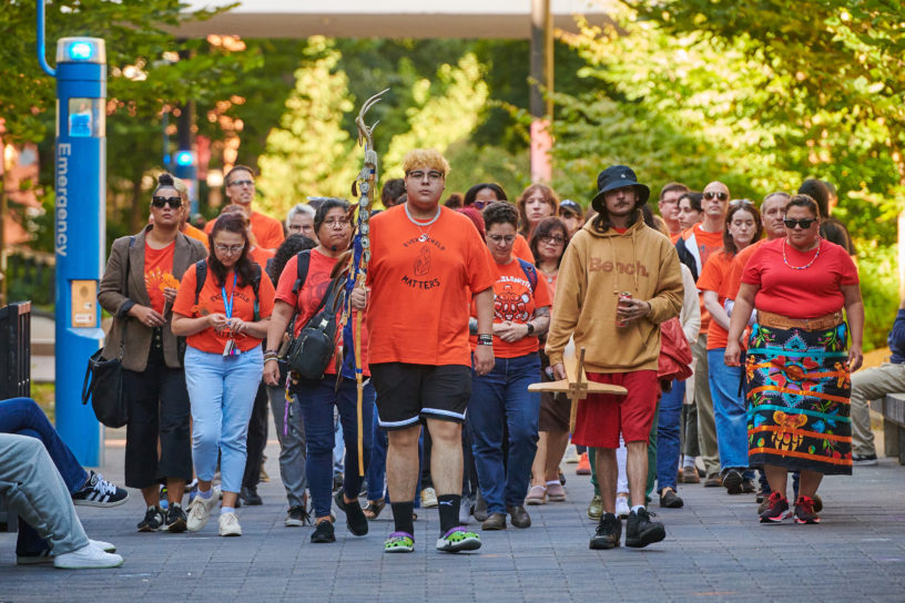 A group of people in orange shirts walk mournfuly down the TMU campus.