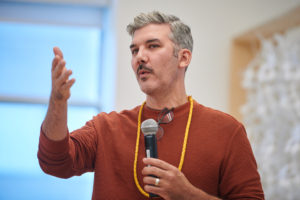 A man in an orange shirt speaks into a mic.