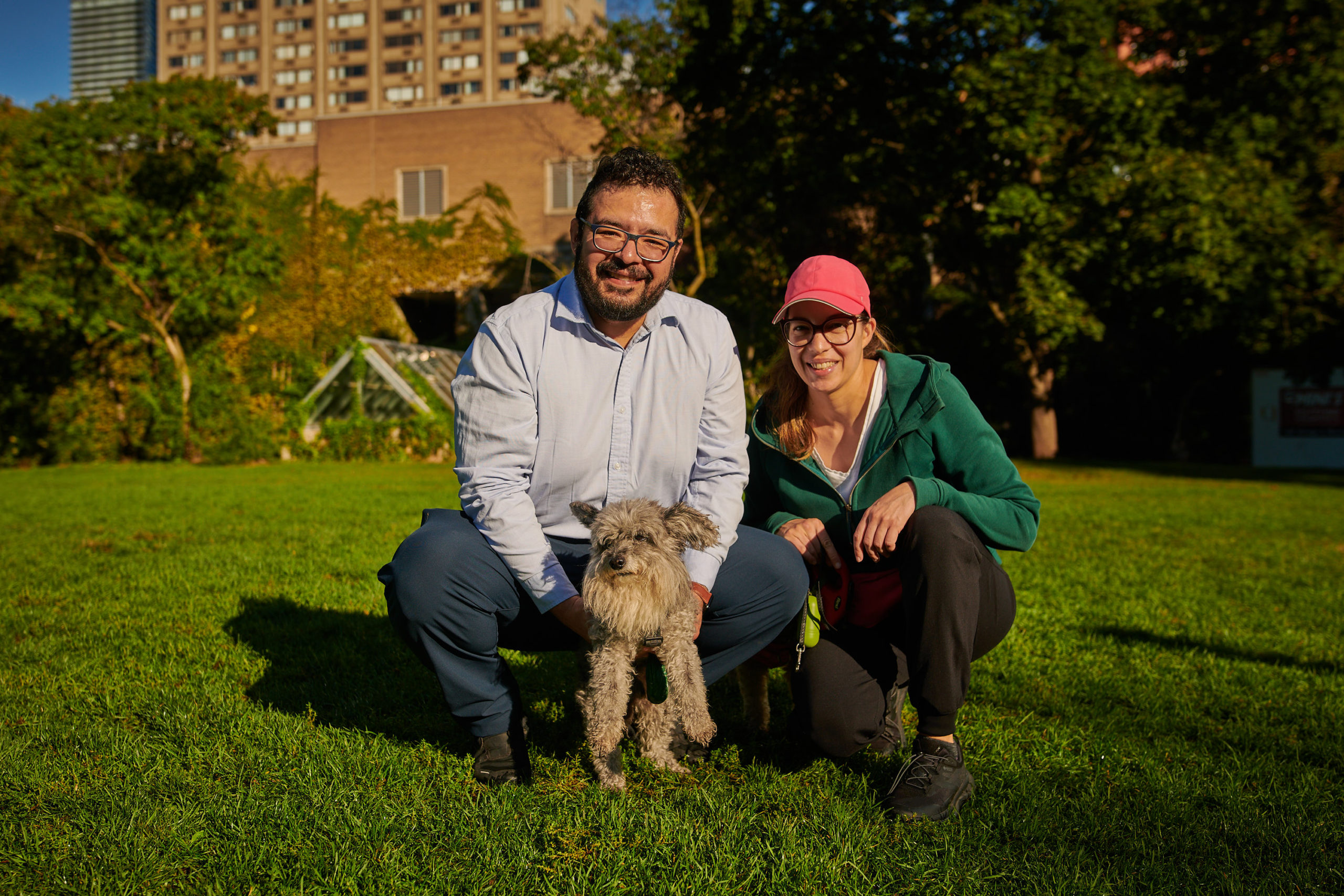 Two dog owners bend down to stand beside their dog in a park field, smiling.