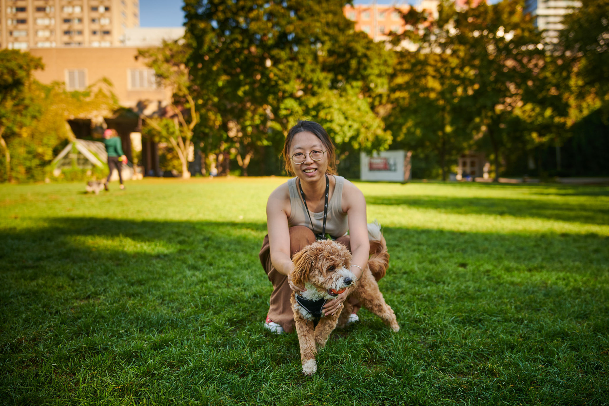 A dog owner bends down to stand beside their excited pet in a park field, smiling towards the camera.