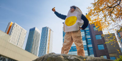 A man wearing an egg costume holds up a half eaten banana.