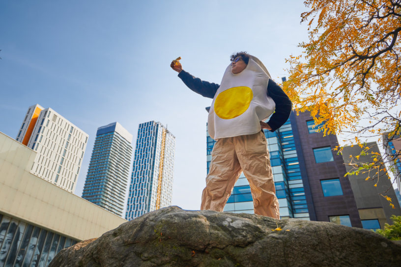 A man wearing an egg costume holds up a half eaten banana.