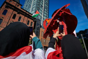 People hold up phones to capture a man that climbed on top of a statue to wave a palestinian flag.