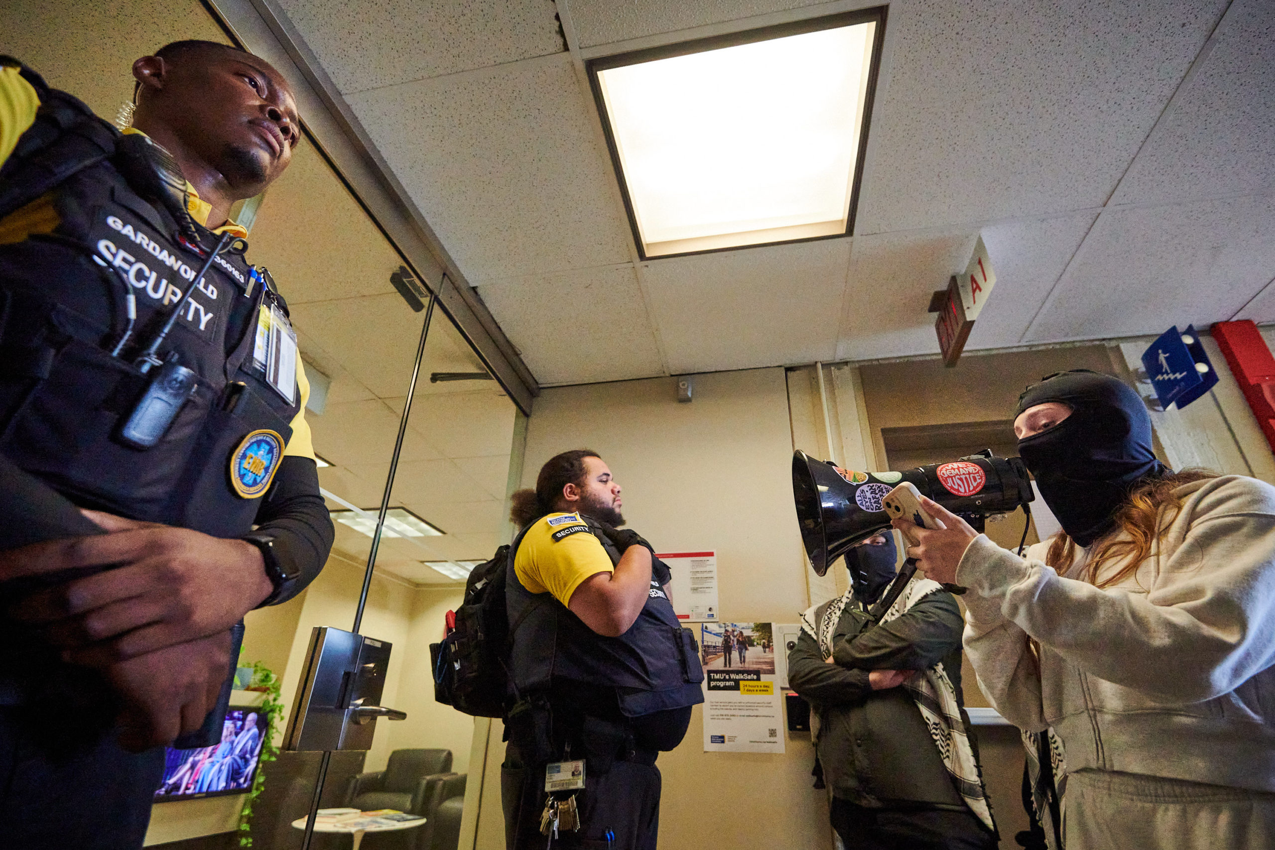 demonstrators articulate demands through megaphone at glass door flanked by two security guards