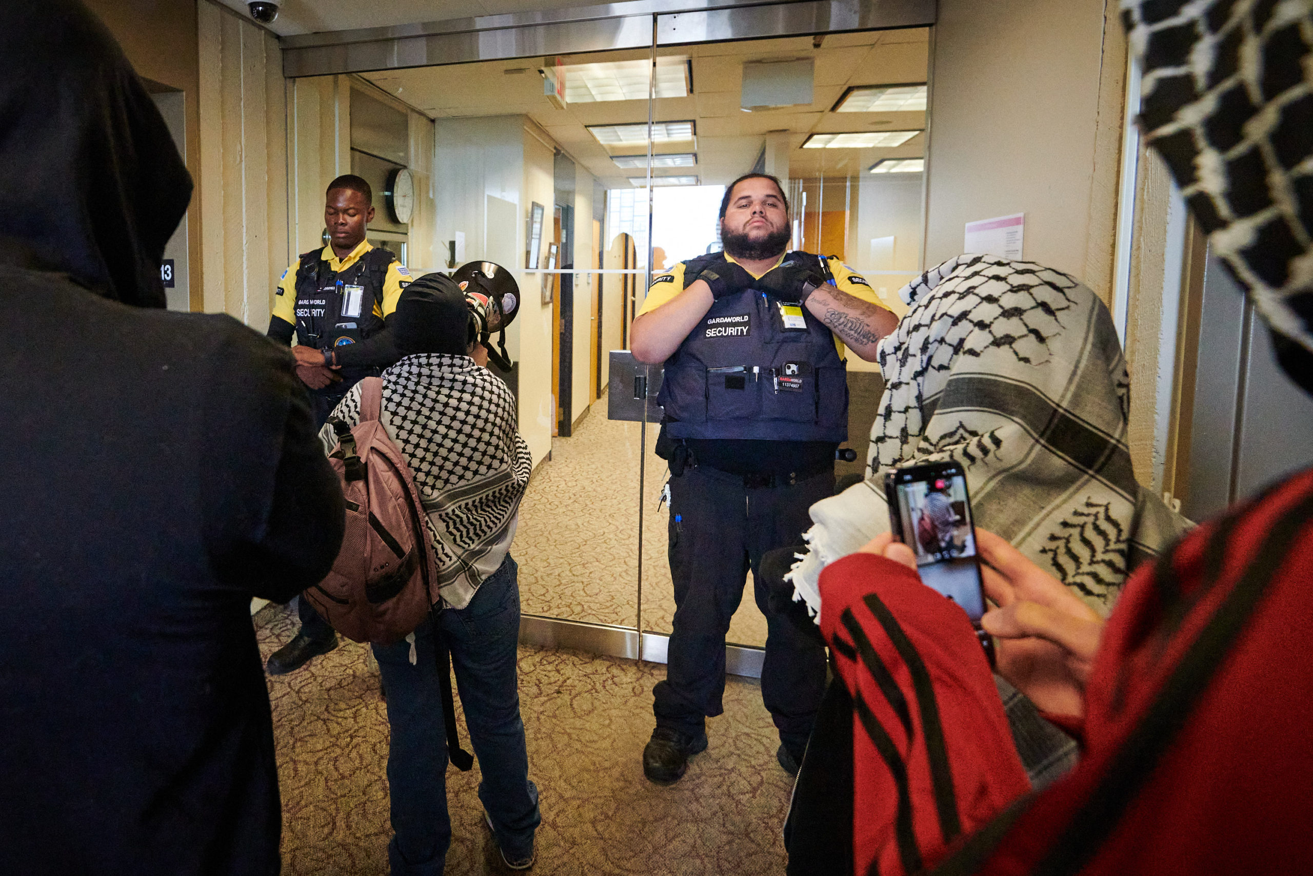 demonstrators articulate demands through megaphone at glass door flanked by two security guards