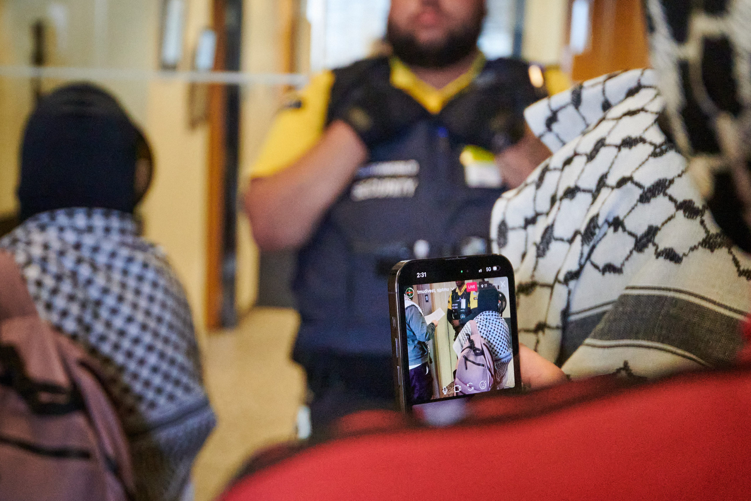 demonstrators film and articulate demands through megaphone at glass door flanked by two security guards