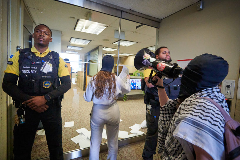 demonstrators articulate demands through megaphone and put letters through door flanked by two security guards