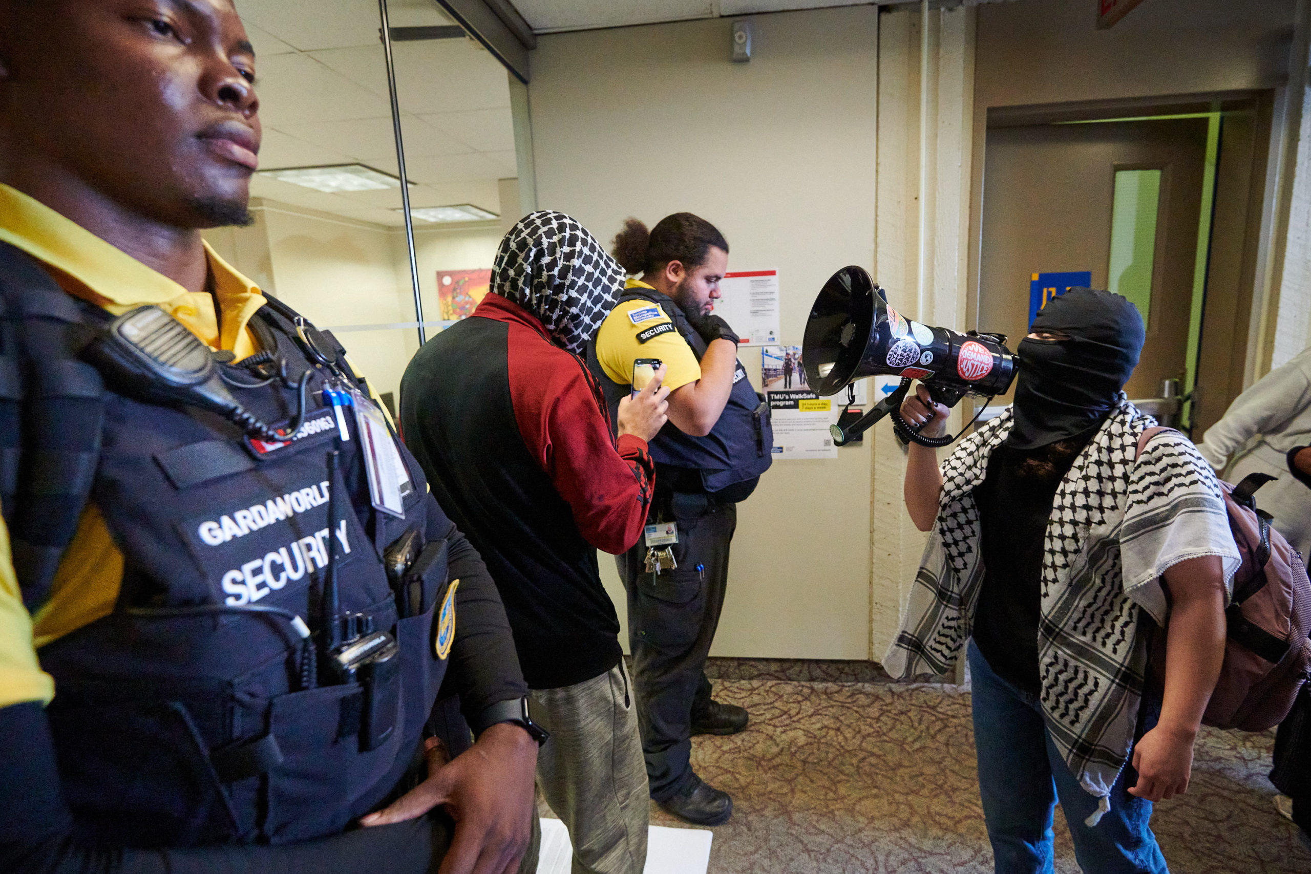 demonstrators articulate demands through megaphone at glass door flanked by two security guards