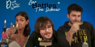 A group of three people posing against a vibrant blue background, each holding a cocktail glass with a light-colored drink garnished with a lemon peel. The text "Drink of the Week" and "Bar Battles: The Sidecar" is displayed at the top. The woman on the left has tanned skin, dark hair, a light top, and bright green nails, while the white man in the middle has glasses, wavy hair, a dark top, and brown hair. The white man on the right is wearing a black hoodie with the words "We The North" in gold lettering.