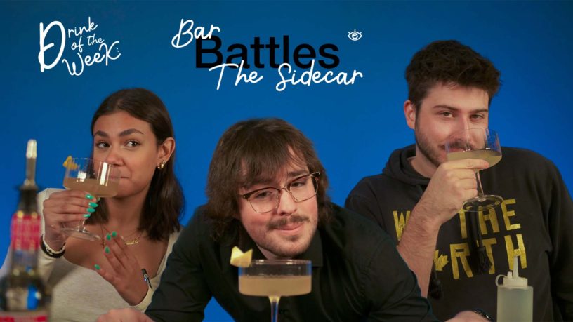 A group of three people posing against a vibrant blue background, each holding a cocktail glass with a light-colored drink garnished with a lemon peel. The text "Drink of the Week" and "Bar Battles: The Sidecar" is displayed at the top. The woman on the left has tanned skin, dark hair, a light top, and bright green nails, while the white man in the middle has glasses, wavy hair, a dark top, and brown hair. The white man on the right is wearing a black hoodie with the words "We The North" in gold lettering.