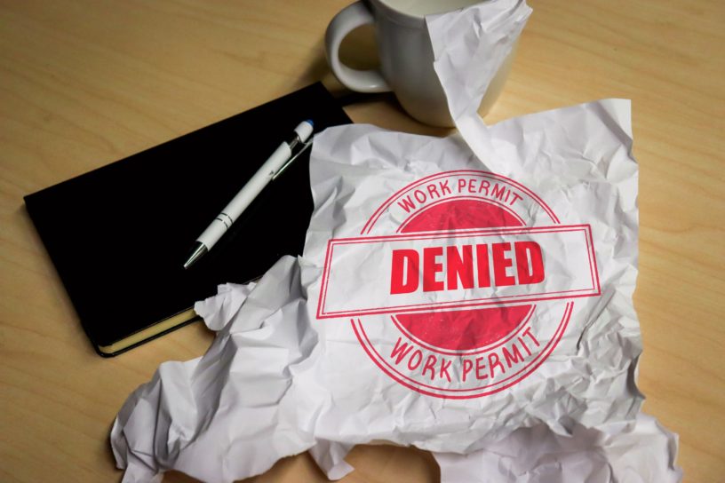 A red stamp reading "DENIED" sits a crumpled piece of paper on a wooden desk.