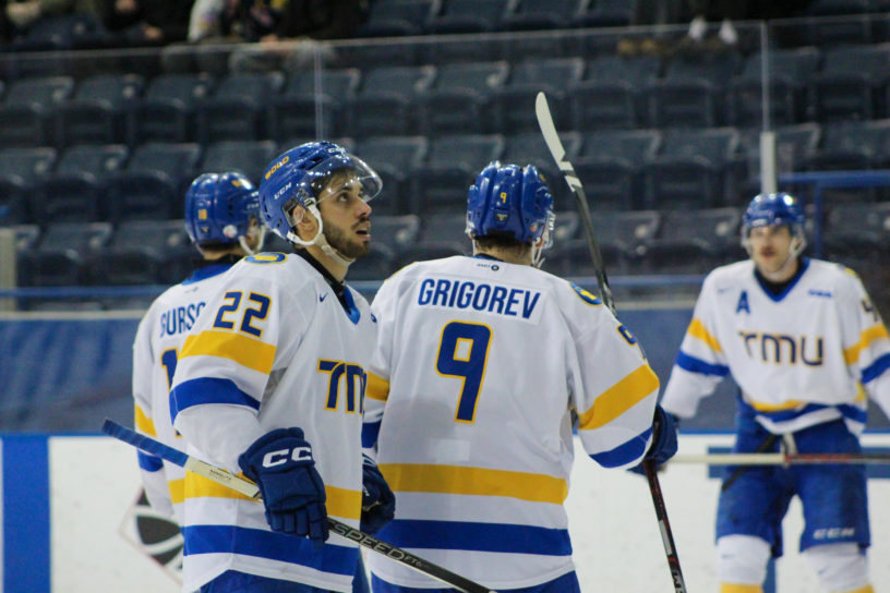 Players on the TMU Bold men's hockey team gather and look up at the scoreboard