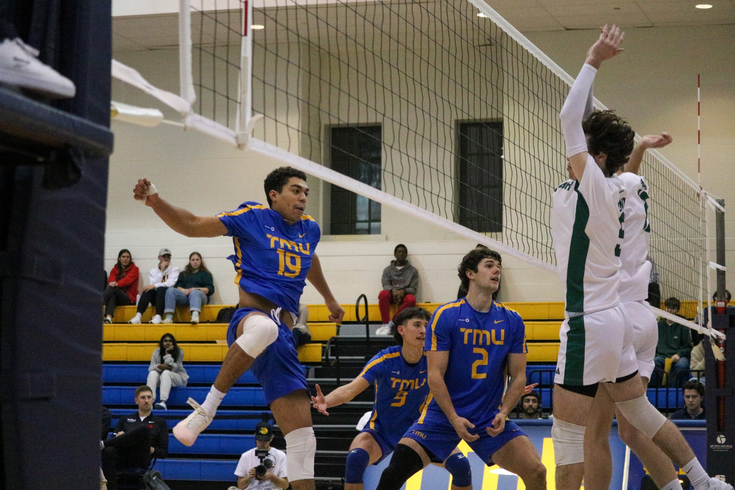 Players on the TMU Bold including Jacob Walker land from jumps to hit a ball over the net towards Trent Excalbur players