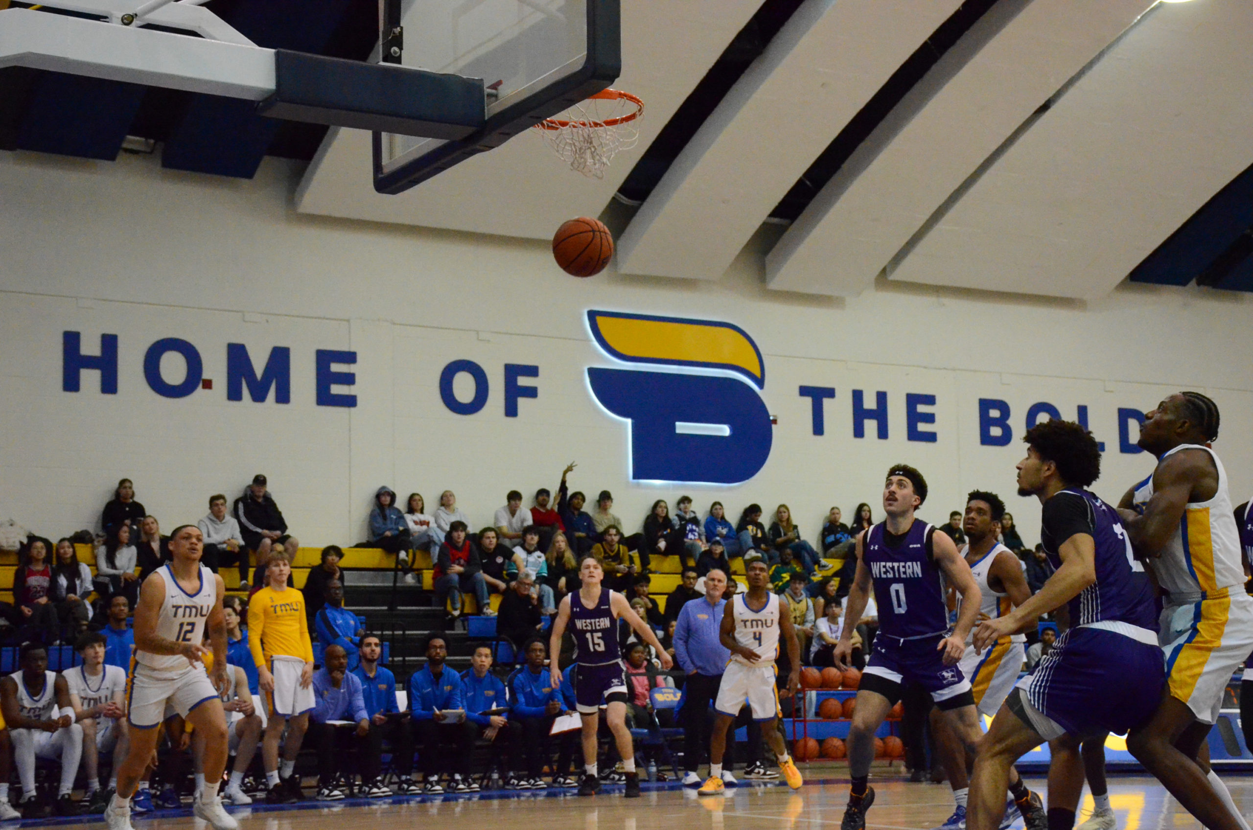 A basketball falls through a net as players from two teams watch in front of a large "Home of the Bold" on the wall