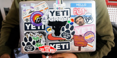 A student in a green sweater holds their laptop, decorated in stickers, close to their chest.