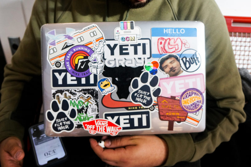 A student in a green sweater holds their laptop, decorated in stickers, close to their chest.