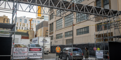 Construction site entrance with warning signs, a gated fence, scaffolding overhead, and a worker in a vest near a parked vehicle.