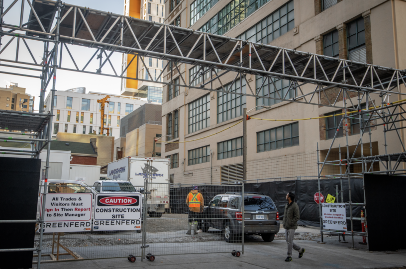 Construction site entrance with warning signs, a gated fence, scaffolding overhead, and a worker in a vest near a parked vehicle.