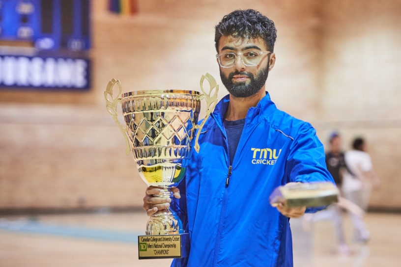 Ahsan Sajjad, member of the TMU cricket team holding the trophy for national champions and pointing the camera with a bat