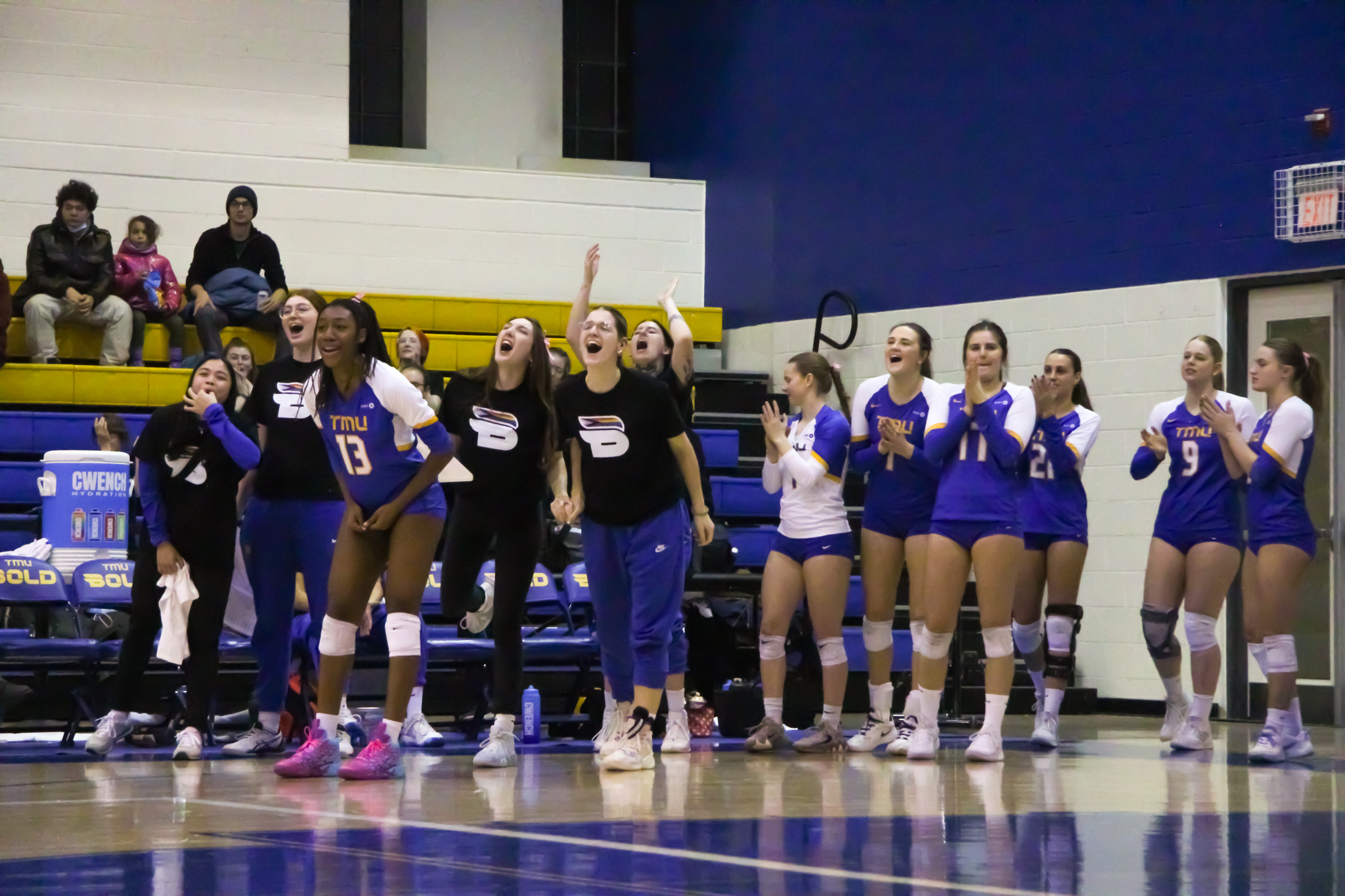 Members of the TMU Bold women's volleyball team cheer and yell from the bench. They are all wearing Pride shirts or Bold jerseys