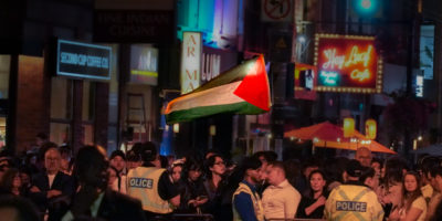 Photo of crowd waiting behind a barricade, with some police standing in front the barricade. A large, bright Palestinian flag is waving in the background of the crowd.