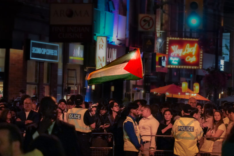 Photo of crowd waiting behind a barricade, with some police standing in front the barricade. A large, bright Palestinian flag is waving in the background of the crowd.