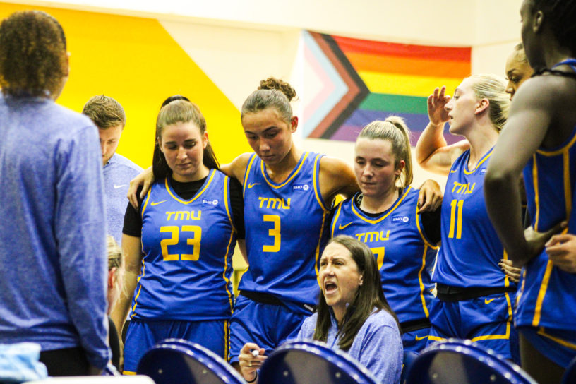 The TMU Bold women's basketball team huddles around head coach Carly Clarke