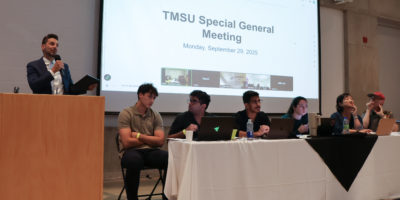 Photo of six people sitting at a white table in front of a powerpoint on a large screen that says "TMSU Special General Meeting."