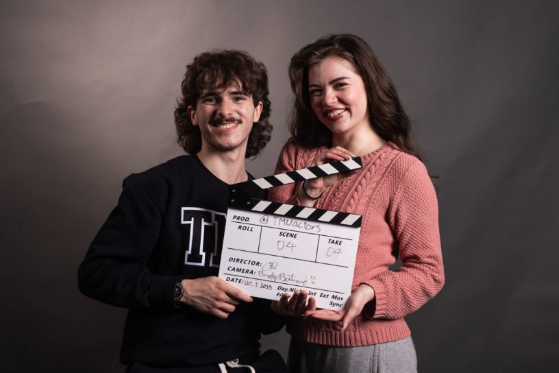Two people look into the camera smiling while they hold a clapperboard.