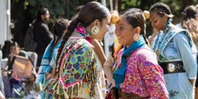 Two smiling young girls standing close to each other, with a potato held up by being wedged between both of their foreheads.