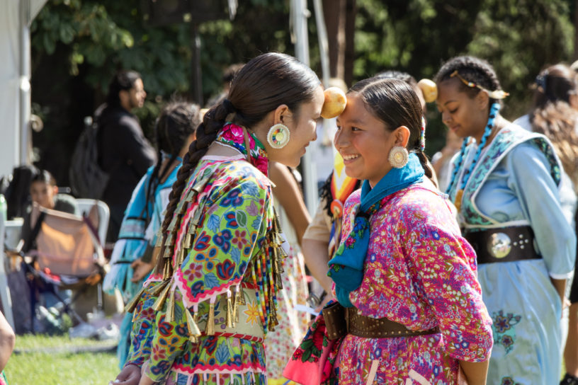 Two smiling young girls standing close to each other, with a potato held up by being wedged between both of their foreheads.