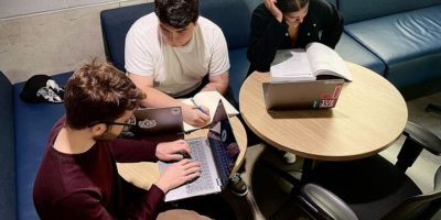 Three students sitting at a table studying with laptops and books open.