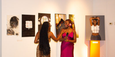Person with long hair wearing a pink dress stands in a room holding a microphone surrounded by art on the wall speaks to another person with long hair in a brown dress.
