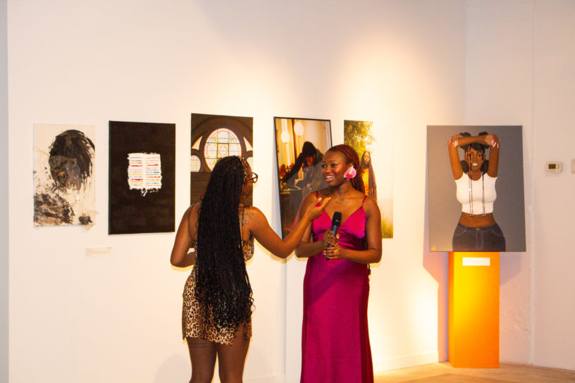 Person with long hair wearing a pink dress stands in a room holding a microphone surrounded by art on the wall speaks to another person with long hair in a brown dress.