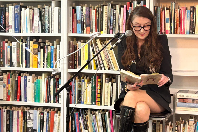 Image of a person looking down at a book reading into a microphone, sitting in front of a bookshelf.