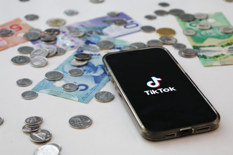 A phone sits on a desk with the TikTok app open, surrounded by bills and coins.