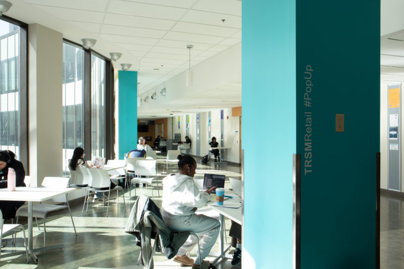 Image of people studying at tables in a building with text on a blue pillar that reads "TRSMRetail #PopUp."