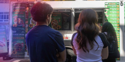 Photo of two people’s backs standing in front of a food truck with a blue and red hue coming from the upper left side of the frame. The blue and red hues cast coloured shadows on the two people.