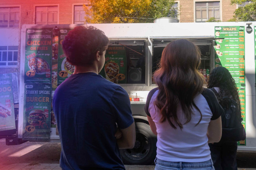 Photo of two people’s backs standing in front of a food truck with a blue and red hue coming from the upper left side of the frame. The blue and red hues cast coloured shadows on the two people.