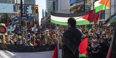 The back of a person speaking to a larger crowd with Palestinian flags in the background