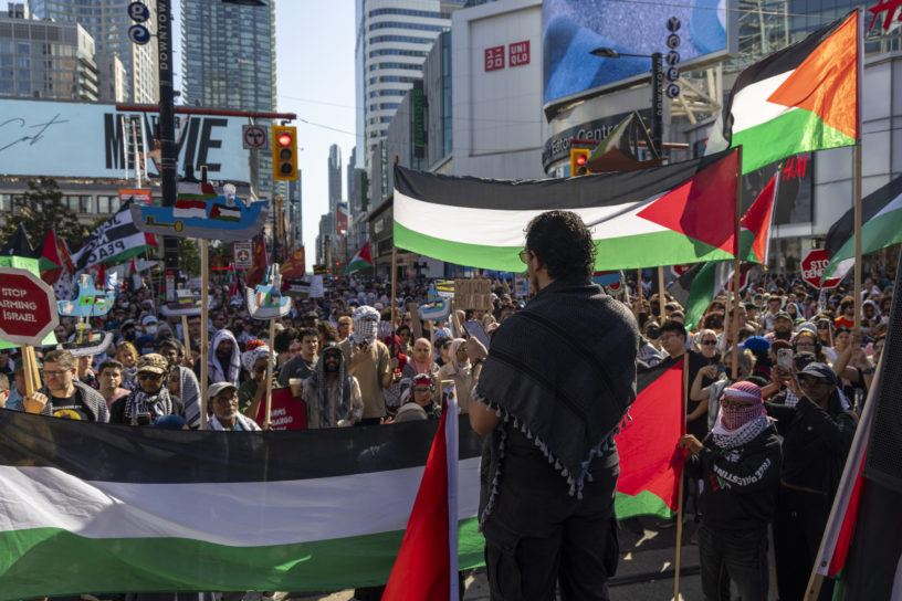 The back of a person speaking to a larger crowd with Palestinian flags in the background