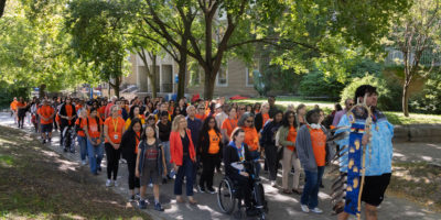 A group of people walking straight, mostly wearing orange shirts down a cement path surrounded by trees.