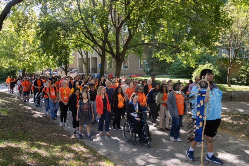 A group of people walking straight, mostly wearing orange shirts down a cement path surrounded by trees.