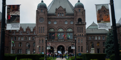 A photo of the Ontario Legislative Assembly, also known as Queen's Park in Toronto with people with placards and banners in the foreground
