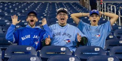 Image of three people in Blue Jays jerseys surrounded by empty blue seats. The people are surprised and excited.