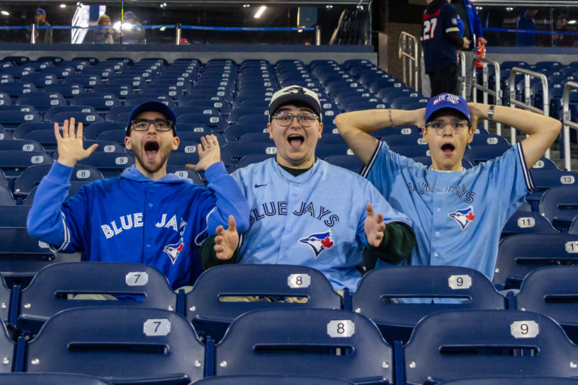 Image of three people in Blue Jays jerseys surrounded by empty blue seats. The people are surprised and excited.