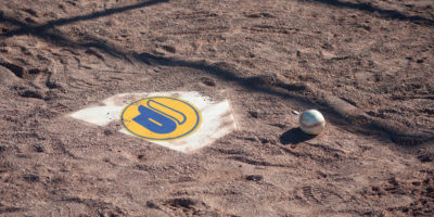 A baseball sits on gravel next to a home plate with a yellow and blue logo on it.